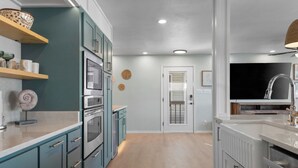 A modern kitchen with teal cabinetry, stainless steel appliances, and a wooden floor.