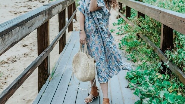 Un pasillo de madera que lleva a una playa, con una persona vestida con un vestido floral y un sombrero.