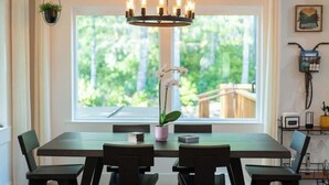 Uma área de refeições com mesa e cadeiras, um lustre, uma planta em vaso e uma janela com vista para vegetação.