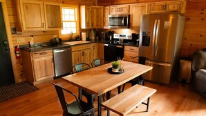 A wooden cabin interior with a kitchen featuring wooden cabinets, a stainless steel refrigerator, and a dining table with chairs. - Treetop Escape | Hot Tub, Hiking, Movie Theater & 5 Private Acres! (Logan)
