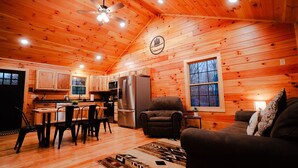 A cozy cabin interior with wooden walls, a dining area, a kitchen with modern appliances, and a comfortable seating area. - Treetop Escape | Hot Tub, Hiking, Movie Theater & 5 Private Acres! (Logan)