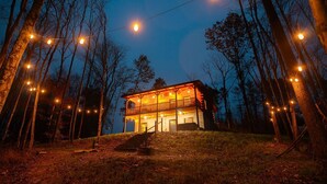 A two-story log cabin with a balcony, surrounded by trees and illuminated by string lights. - Treetop Escape | Hot Tub, Hiking, Movie Theater & 5 Private Acres! (Logan)