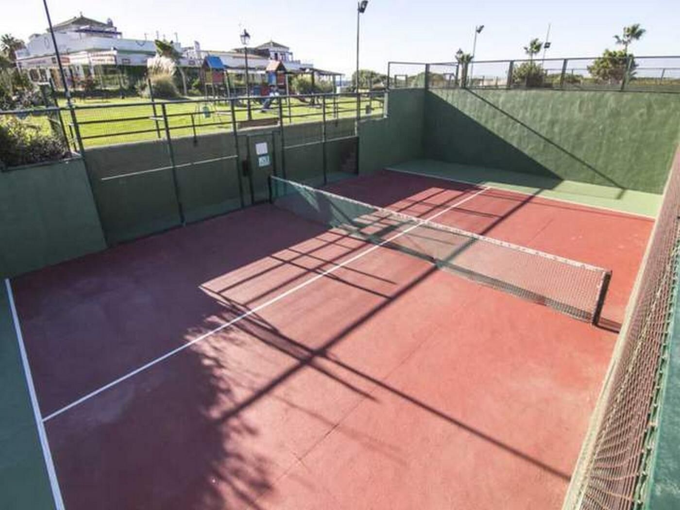 Een indoor tennisbaan met een net, groene wanden en een heldere lucht zichtbaar door de ramen.