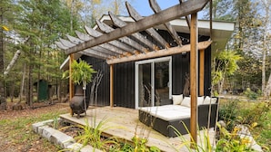 Un terrazzo in legno con una pergola, un edificio nero con porte in vetro e un’area relax con cuscini.