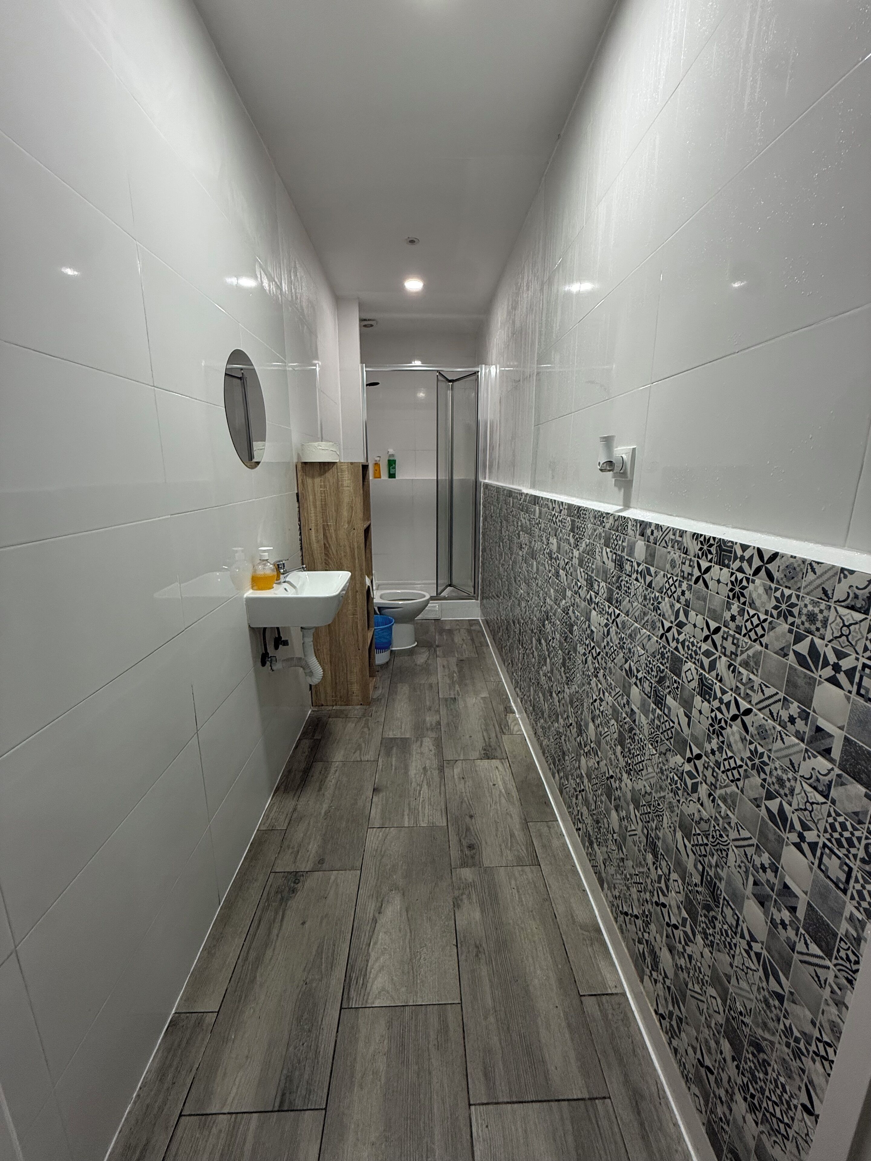 Un baño estrecho con paredes de azulejos blancos, un tocador de madera, un lavabo y una zona de ducha.