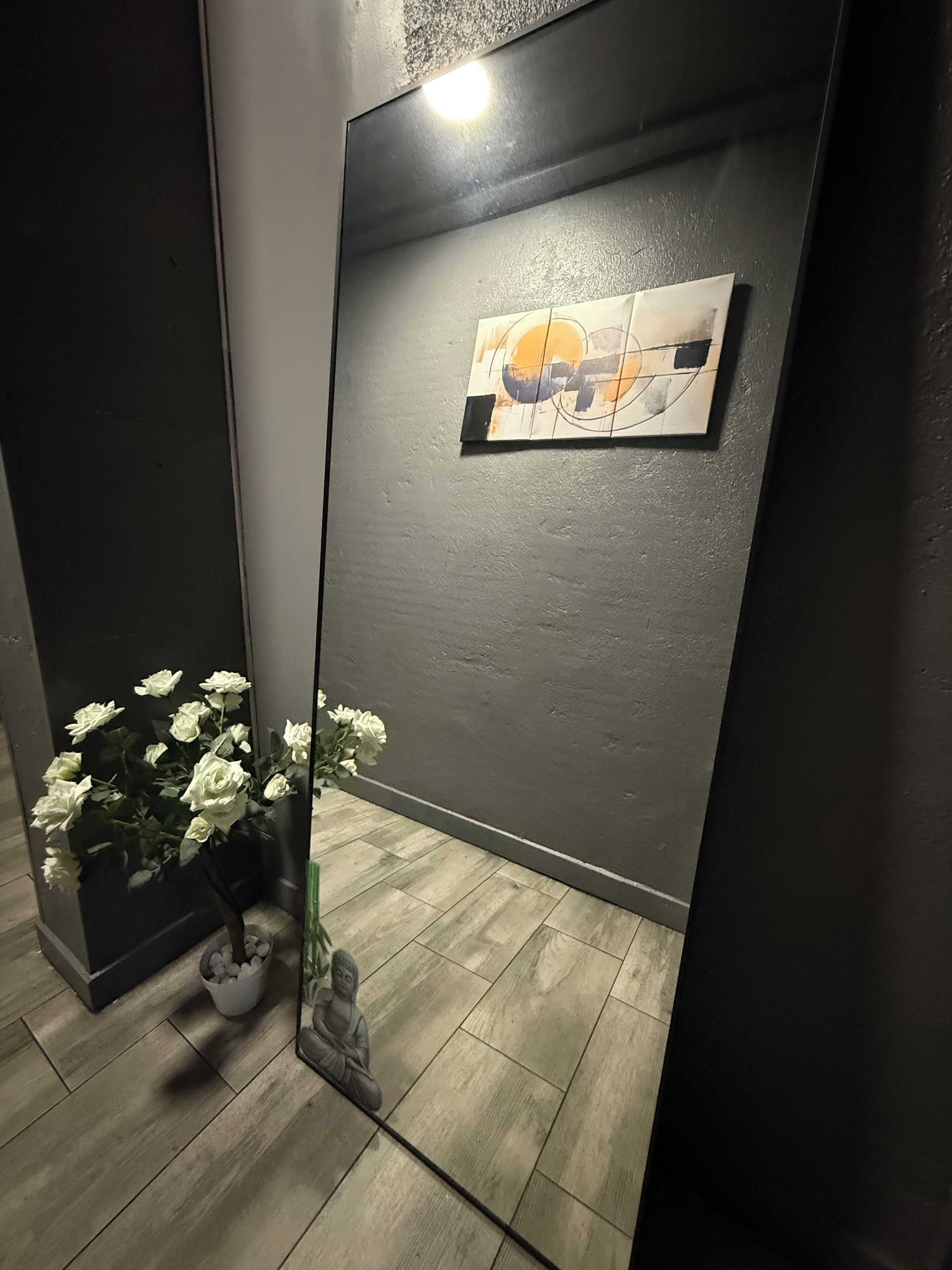 A modern room with a large mirror reflecting a white flower arrangement and a statue. A painting is visible on the wall, and the floor is tiled.