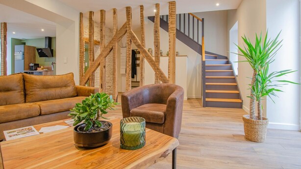 Una sala de estar moderna con una mesa de centro de madera, un sofá de cuero marrón y un sillón marrón, además de una planta en maceta. Se aprecia una escalera con barandilla de madera al fondo.