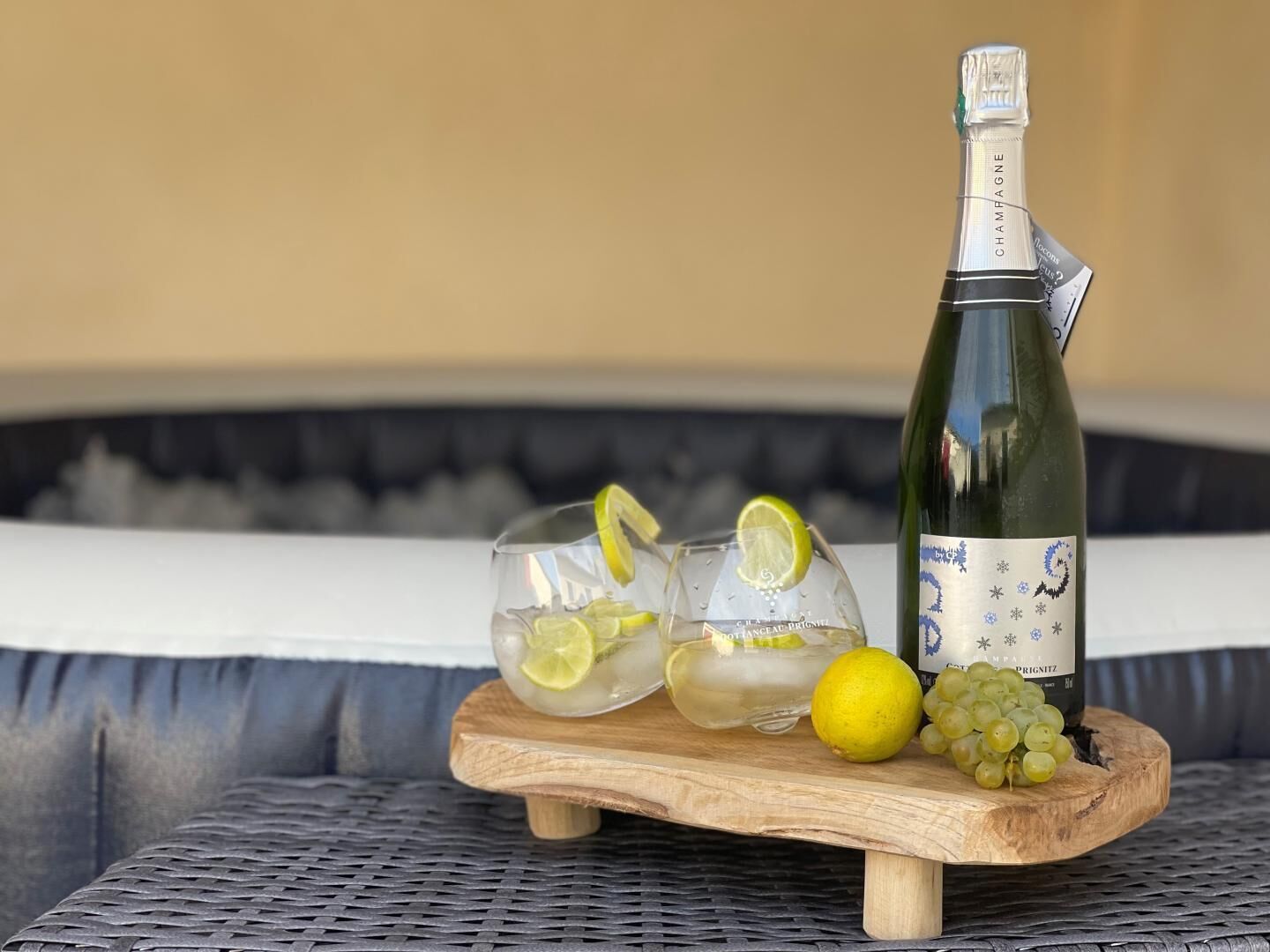 A bottle of champagne, two glasses with lime slices, a lemon, and grapes on a wooden board.