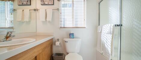 Et badeværelse med et trækommode, toilet, brusekabine i glas og to vinduer med skodder.