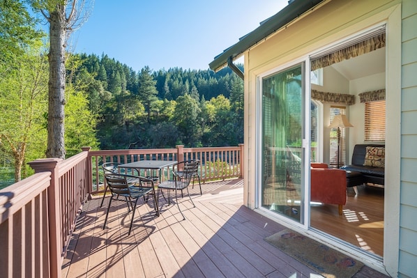 Eine Terrasse mit Tisch und Stühlen, eine Glasschiebetür zu einem Wohnbereich mit Sofa und Lampe sowie ein bewaldeter Hügel im Hintergrund.