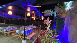 Un espace extérieur aménagé avec des tables recouvertes de nappes à carreaux, des bancs en bois, des lanternes suspendues et des plantes en pot.