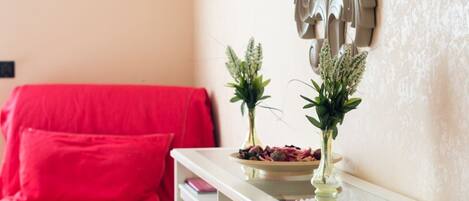 Una sala de estar con un sofá rojo, una mesita auxiliar blanca con un bol de aperitivos, y dos jarrones con plantas.