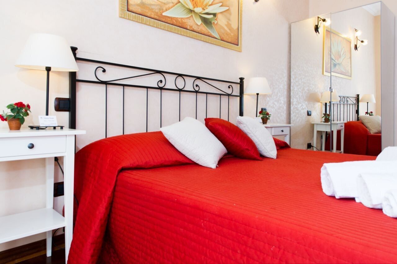 A hotel room with a red bedspread, white pillows, a metal headboard, bedside tables with lamps, a small desk with a plant, and a mirror.