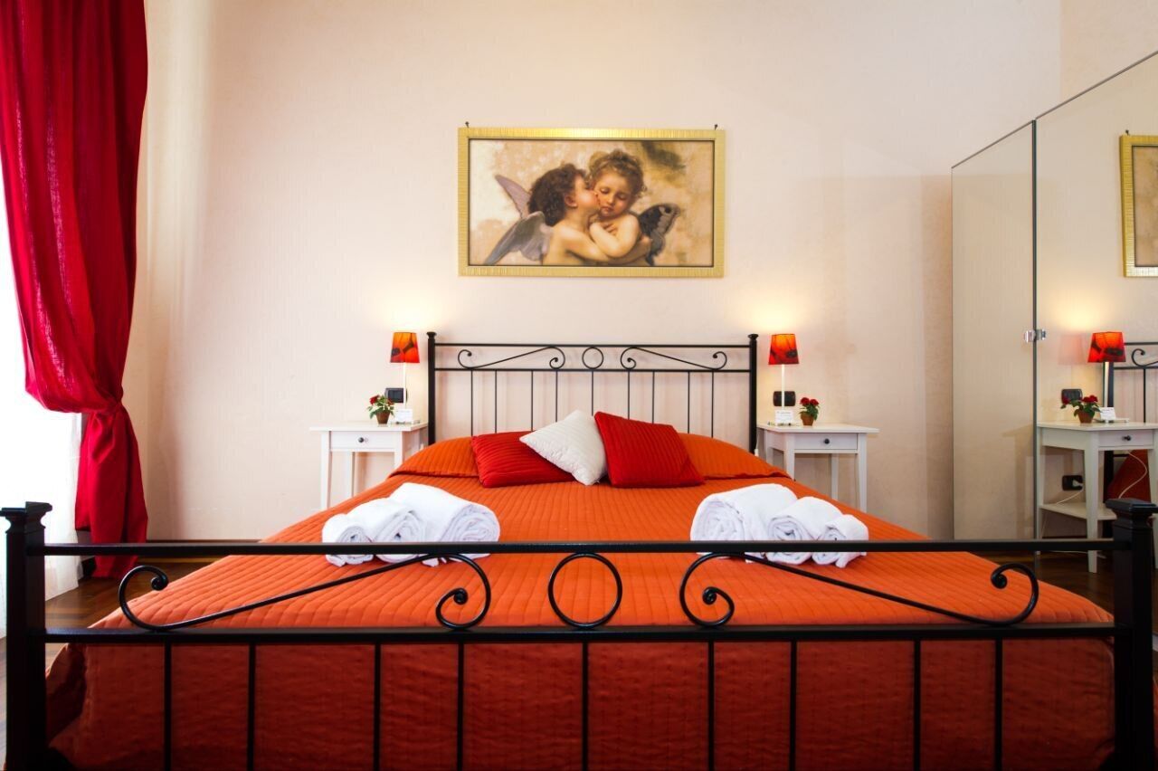A hotel room with a bed, red curtains, a painting of cherubs, two bedside tables with lamps, and a mirror.