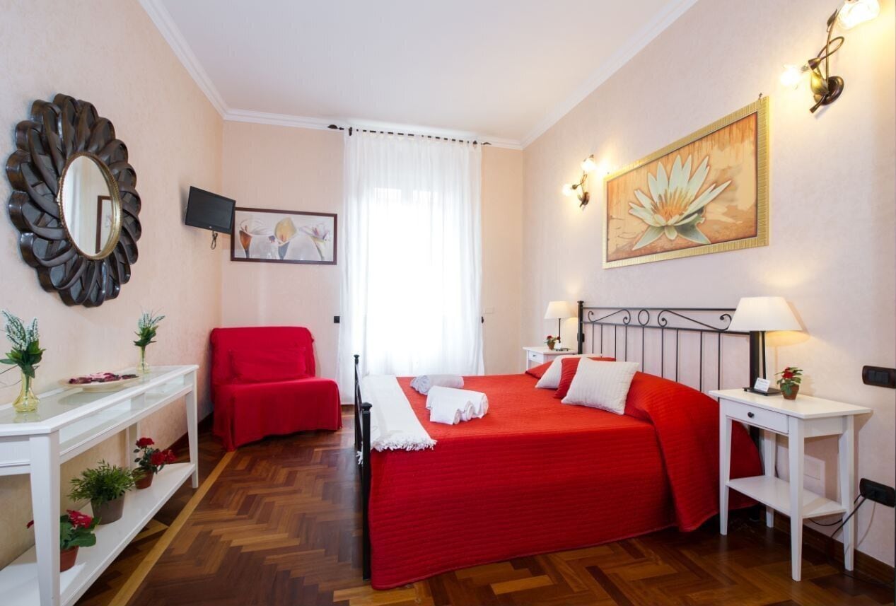 Quarto de hotel com cama vermelha, móveis brancos, televisão, pintura e espelho.