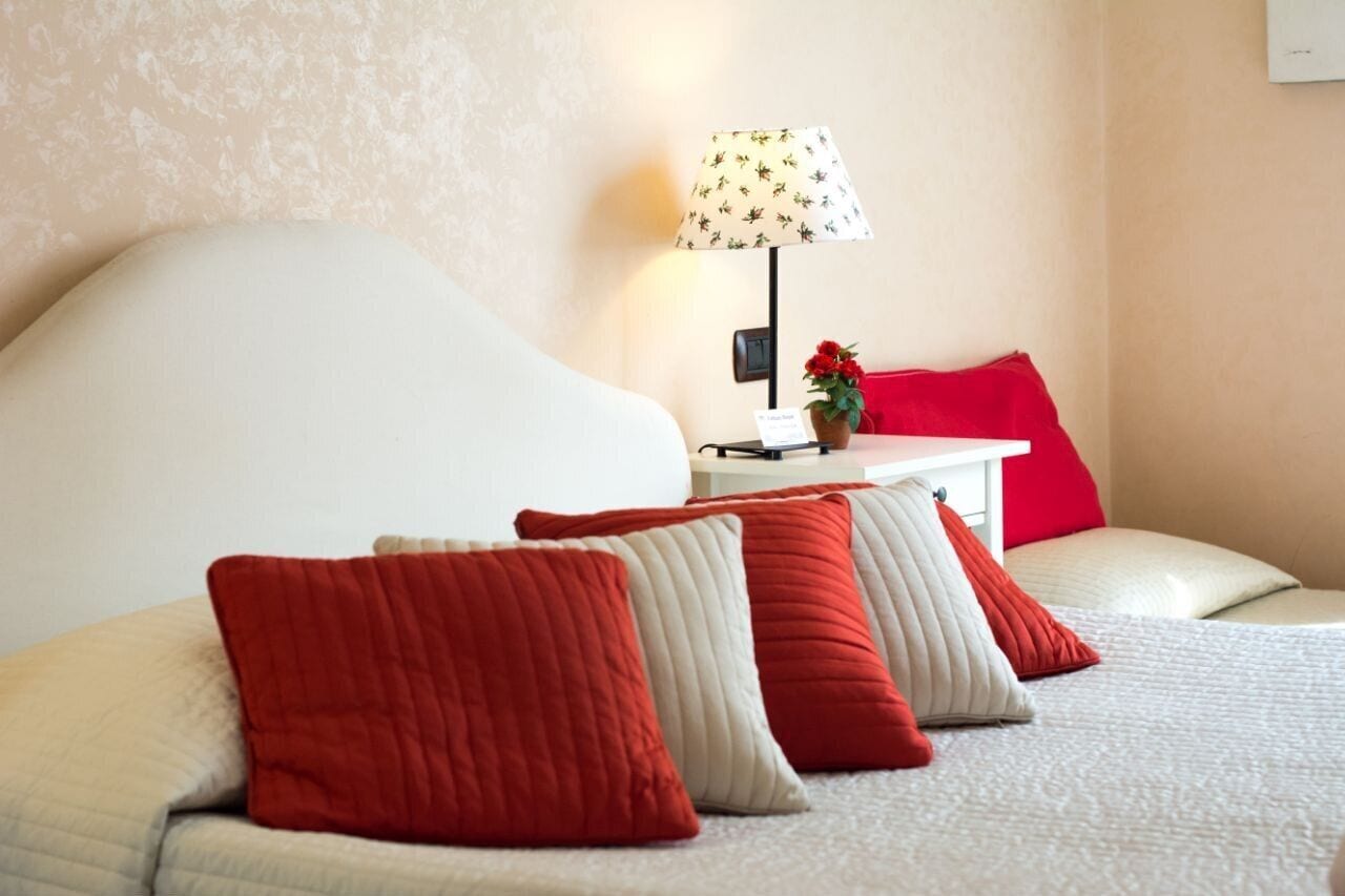 A neatly made bed with red and white pillows, a bedside table with a lamp, and a small red flower vase.