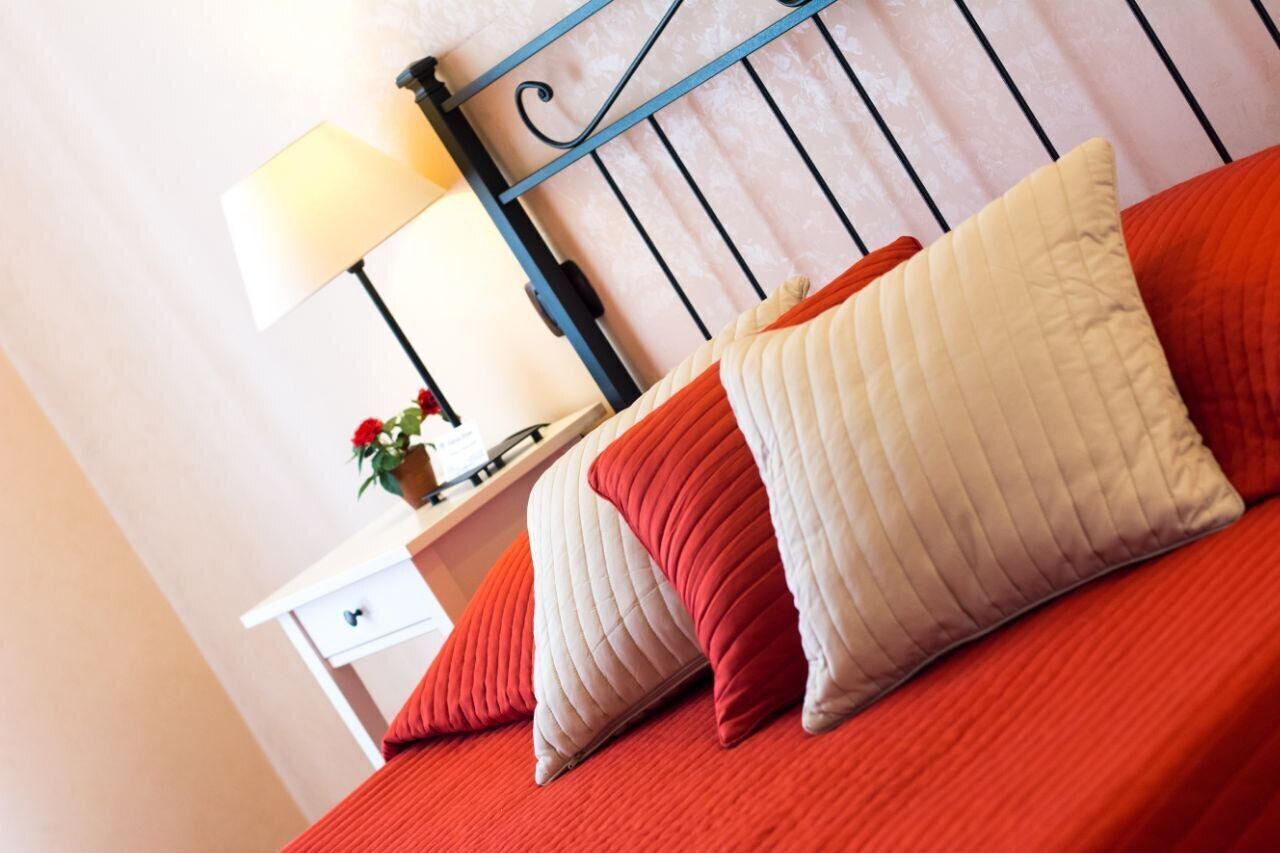 Een bed met rode beddengoed en meerdere kussens, een nachtkastje met een lamp en een vaas bloemen, en een witte nachtkast met een lade.