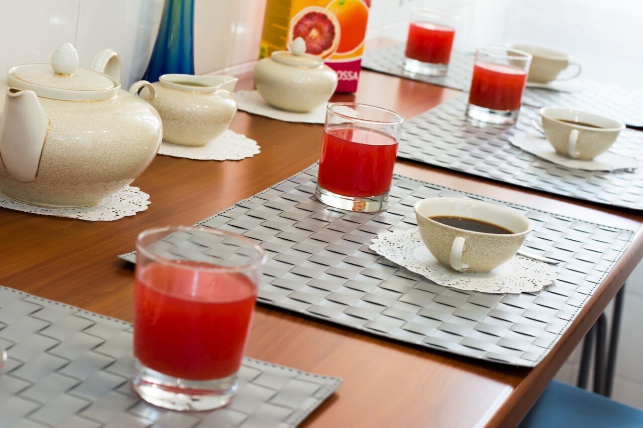 A breakfast setting with teapots, cups, and a glass of red juice on a table with a woven placemat.