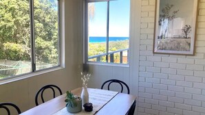 A dining area with a table set for four, large windows offering a view of the sea and greenery, and a framed picture on the wall.
