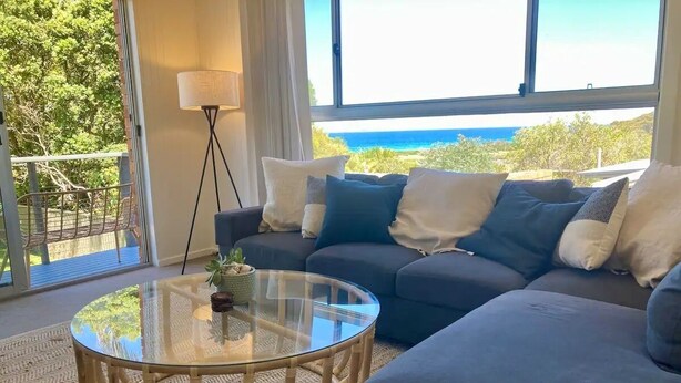 A modern living room with a glass coffee table, dark sofas, and a view of the ocean.