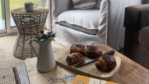 A cozy living room with a grey armchair, a wooden coffee table with croissants, a white vase with a plant, and a lamp.