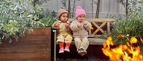 Deux enfants sont assis sur un banc en bois dans un jardin, avec un foyer au premier plan.