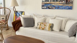 A living room with a white sofa, a wooden coffee table, a wicker floor rug, a lamp, and a painting of a coastal town.