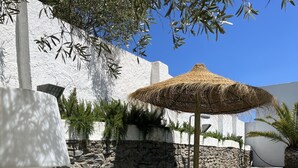 A blue sofa, a wooden table with a plate, and a chair in an outdoor setting with stone walls and a thatched umbrella.