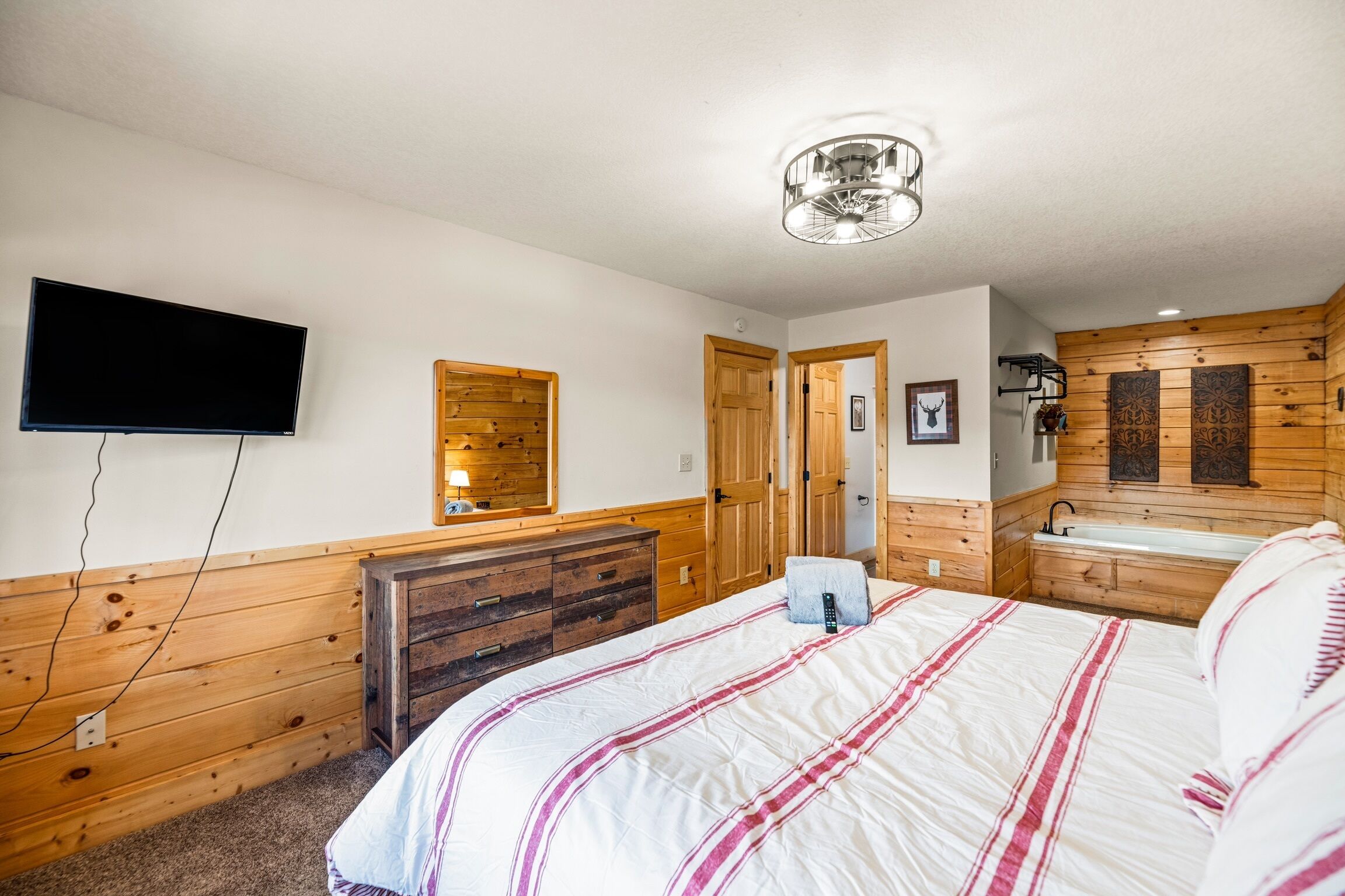 Une chambre à coucher comprenant un lit, une commode en bois, une télévision, ainsi qu’une salle de bain avec un lavabo et un miroir.