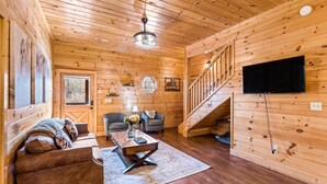 A wooden interior with a sofa, chairs, a coffee table, a TV, and a staircase.