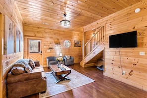 Ein Holzinterieur mit einer Couch, Sesseln, einem Couchtisch, einem Fernseher und einer Treppe.