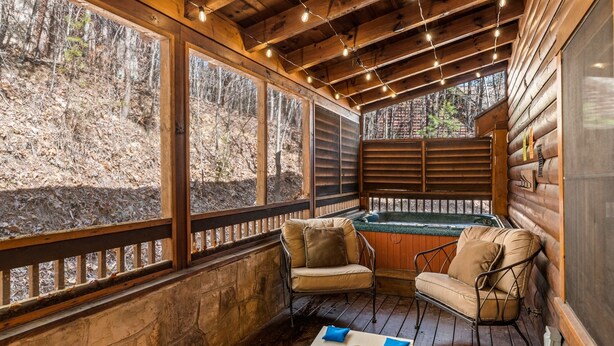 A wooden cabin with a hot tub, two beige cushioned chairs, and a stone wall.