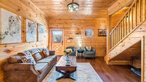 A wooden cabin interior with a sofa, coffee table, and two chairs. A staircase leads to an upper level.