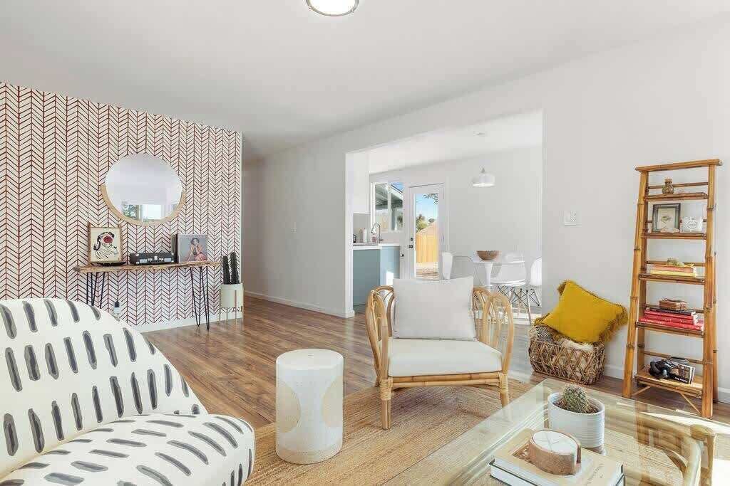 A modern living room with a herringbone patterned wall, a round mirror, a white sofa, a wicker chair, a glass coffee table, and a bookshelf.