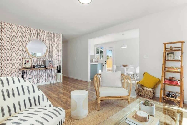 A modern living room with a herringbone patterned wall, a round mirror, a white sofa, a wicker chair, a glass coffee table, and a bookshelf. - Audrey's Place, New Hot Tub, Mins to Joshua Tree (Joshua Tree)