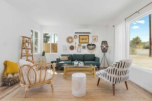 A living room with a green sofa, a coffee table, a wicker chair, a floor lamp, and a large window.