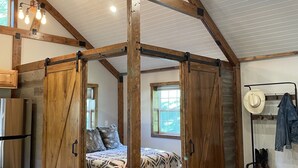 A rustic interior with exposed wooden beams, a bed with patterned bedding, a dining area with a marble countertop, and a coat rack with hats and boots.
