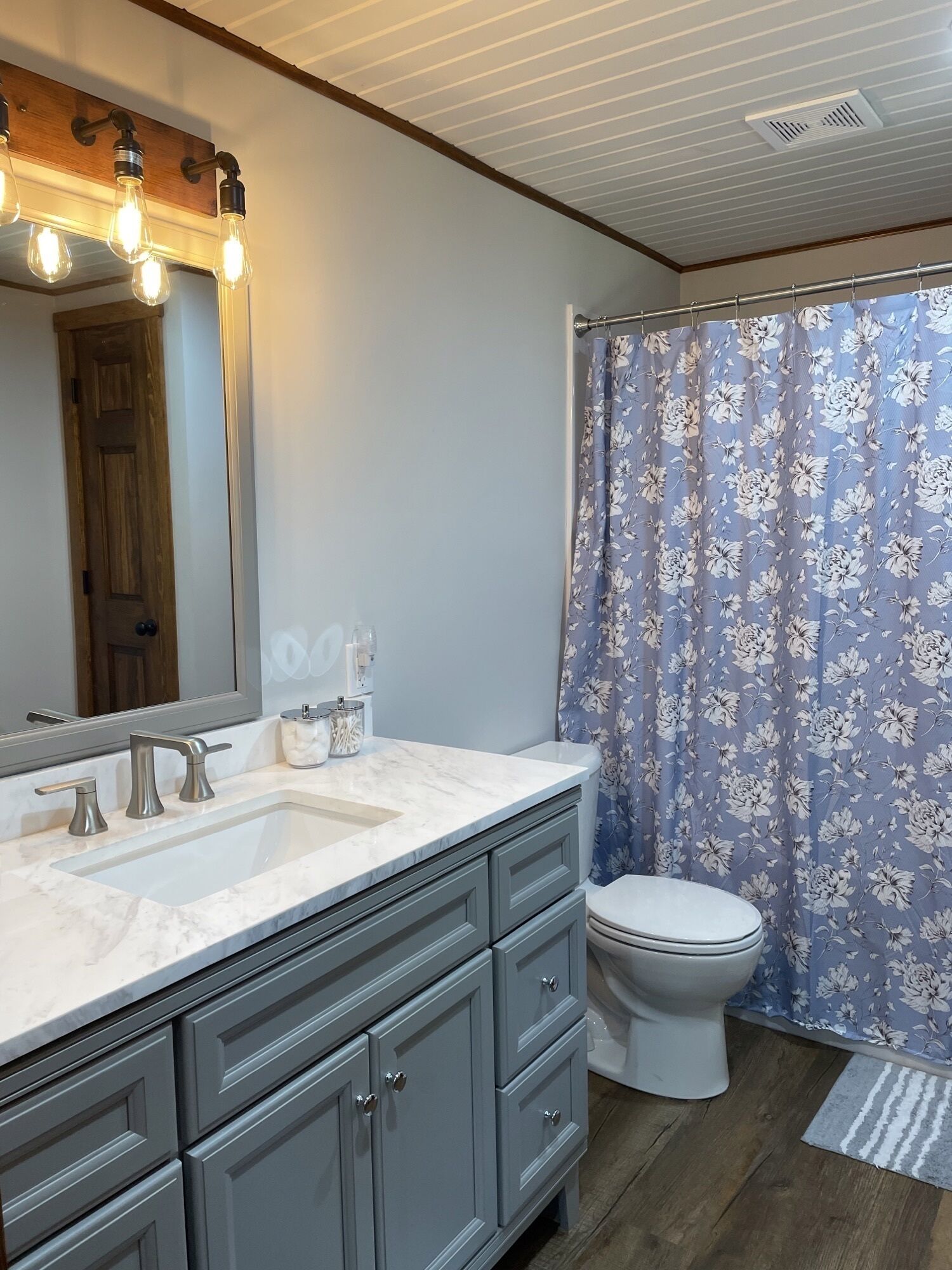 Une salle de bain avec un lavabo blanc, des meubles gris, des toilettes et un rideau de douche à motifs floraux.