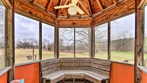 A wooden gazebo with a bench, large windows, and a ceiling fan.