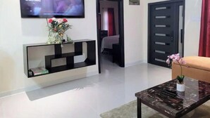 Een moderne woonkamer met een flatscreen televisie, een zwarte console tafel met een bloevas, een glazen salontafel en een deur die naar een andere kamer leidt.