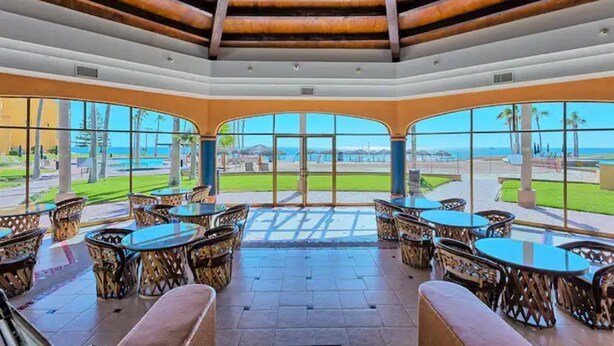 A covered outdoor seating area with tables and chairs, large windows offering a view of the beach and ocean, and a wooden ceiling structure.