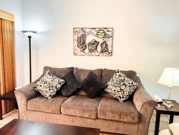 Ein Wohnzimmer mit braunem Sofa, gemusterten Kissen, einem Wandbild, einer Stehlampe und einem kleinen Tisch mit Lampe.