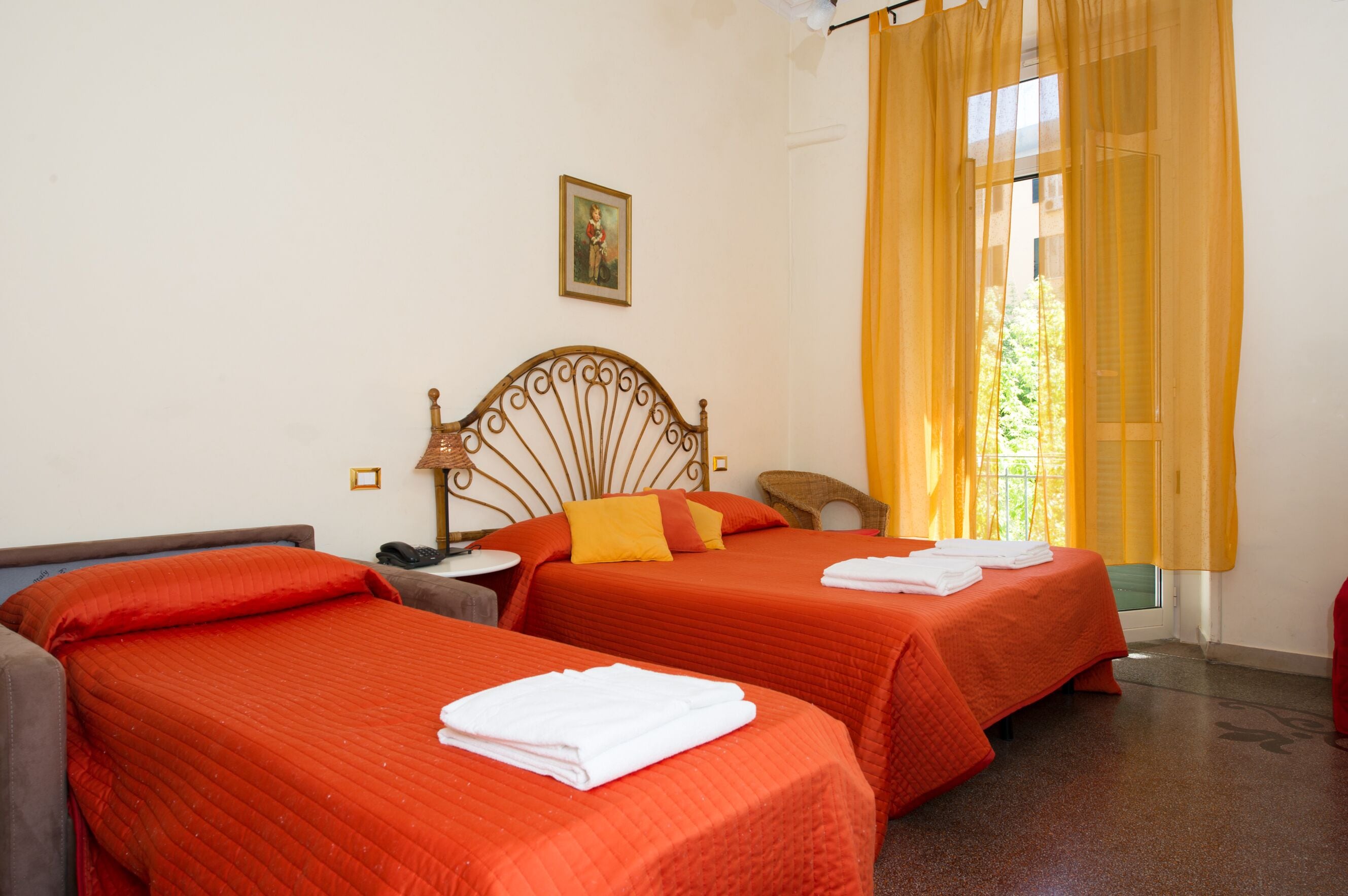 A hotel room with two beds, a single chair, a bedside table, a lamp, and a window with yellow curtains.