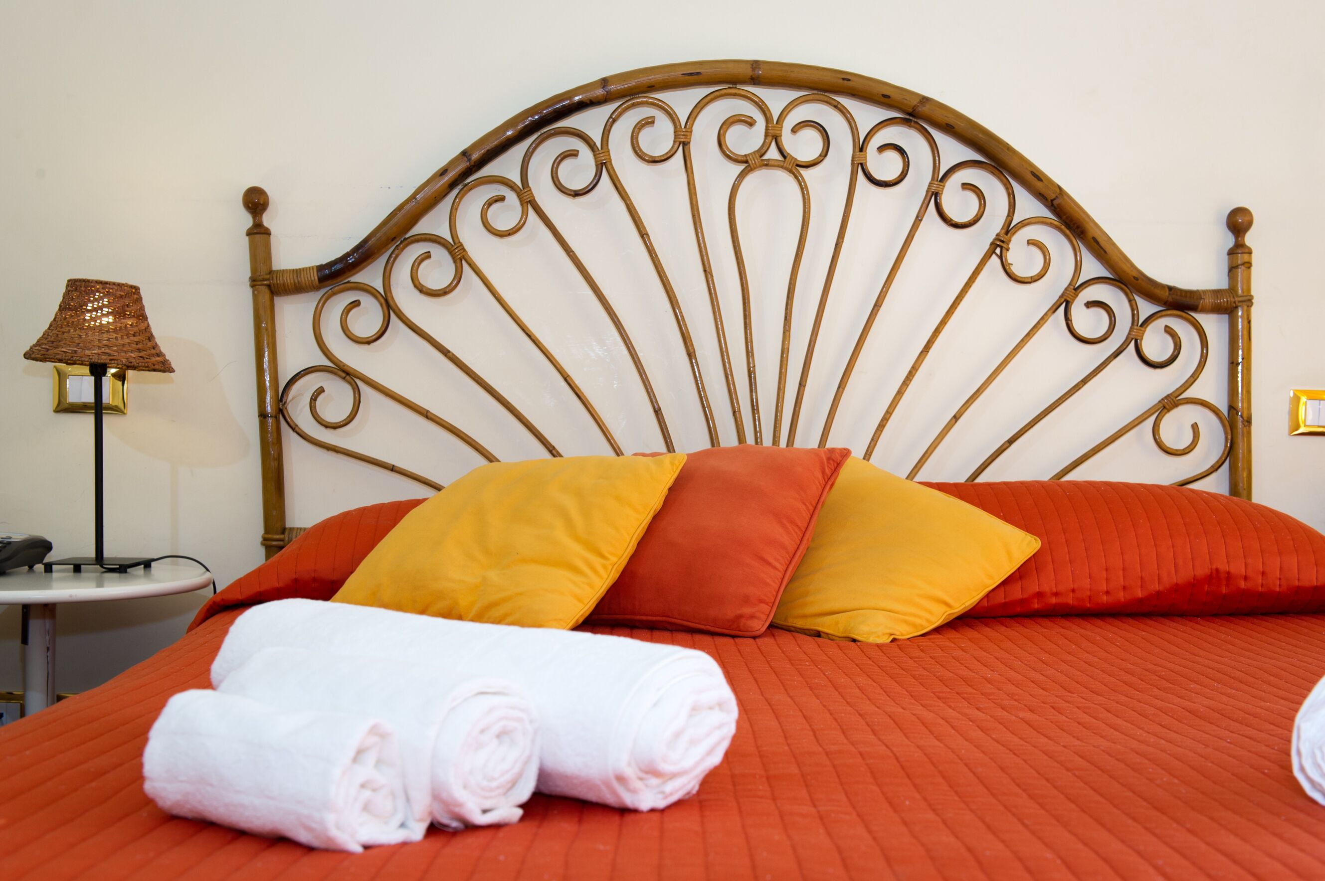 Una cama bien hecha con ropa de cama roja, dos toallas enrolladas y una lámpara sobre una mesita de noche.