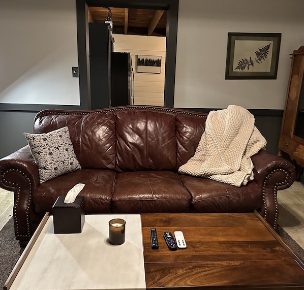 Un sofá de cuero marrón con una manta y almohadones estampados, una mesa blanca con una vela y controles remotos, y un helecho enmarcado en la pared.