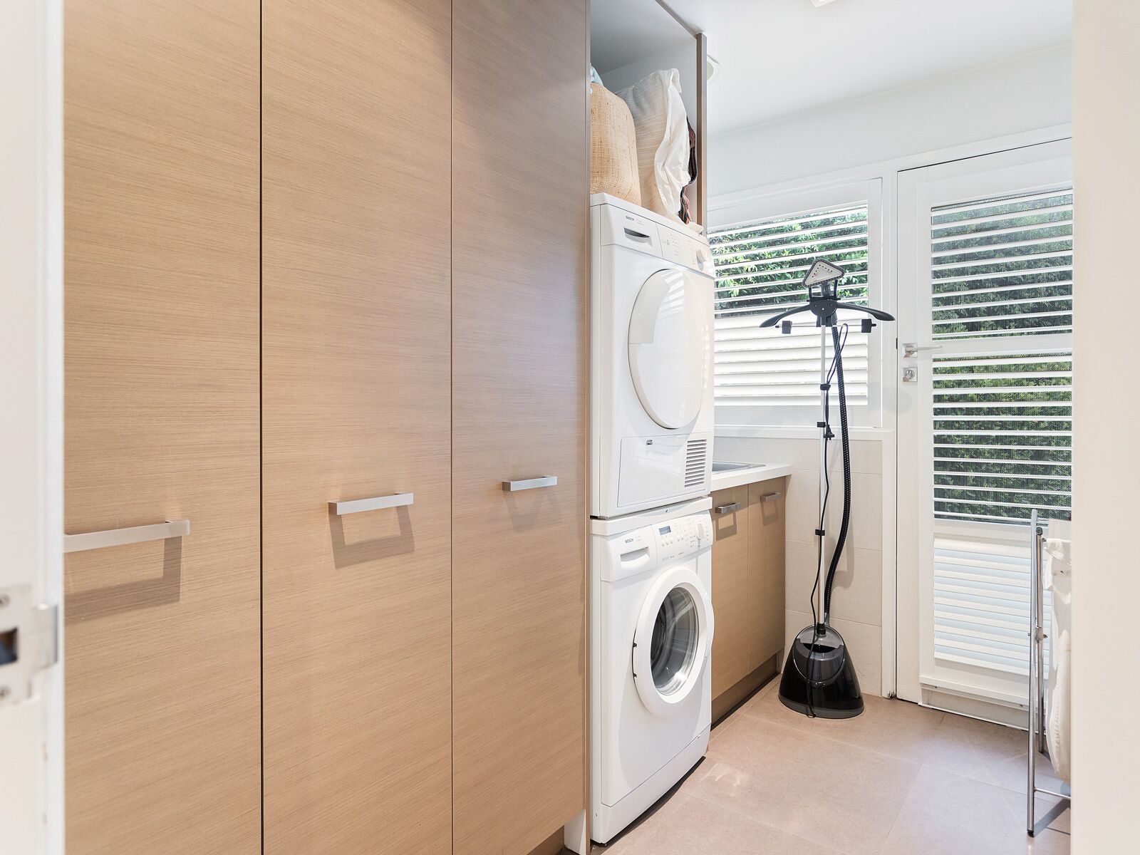 A laundry room with a front-loading washer and dryer, wooden cabinets, and a window with blinds.