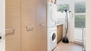 Une buanderie équipée d’un lave-linge et d’un sèche-linge à chargement frontal, de meubles en bois et d’une fenêtre avec des stores.