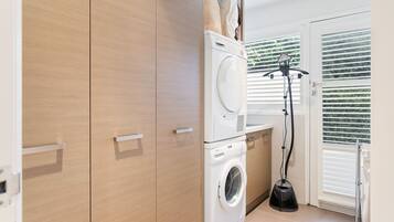 A laundry room with a front-loading washer and dryer, wooden cabinets, and a window with blinds.