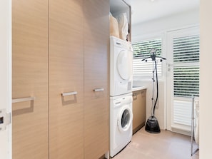 A laundry room with a front-loading washer and dryer, wooden cabinets, and a window with blinds.