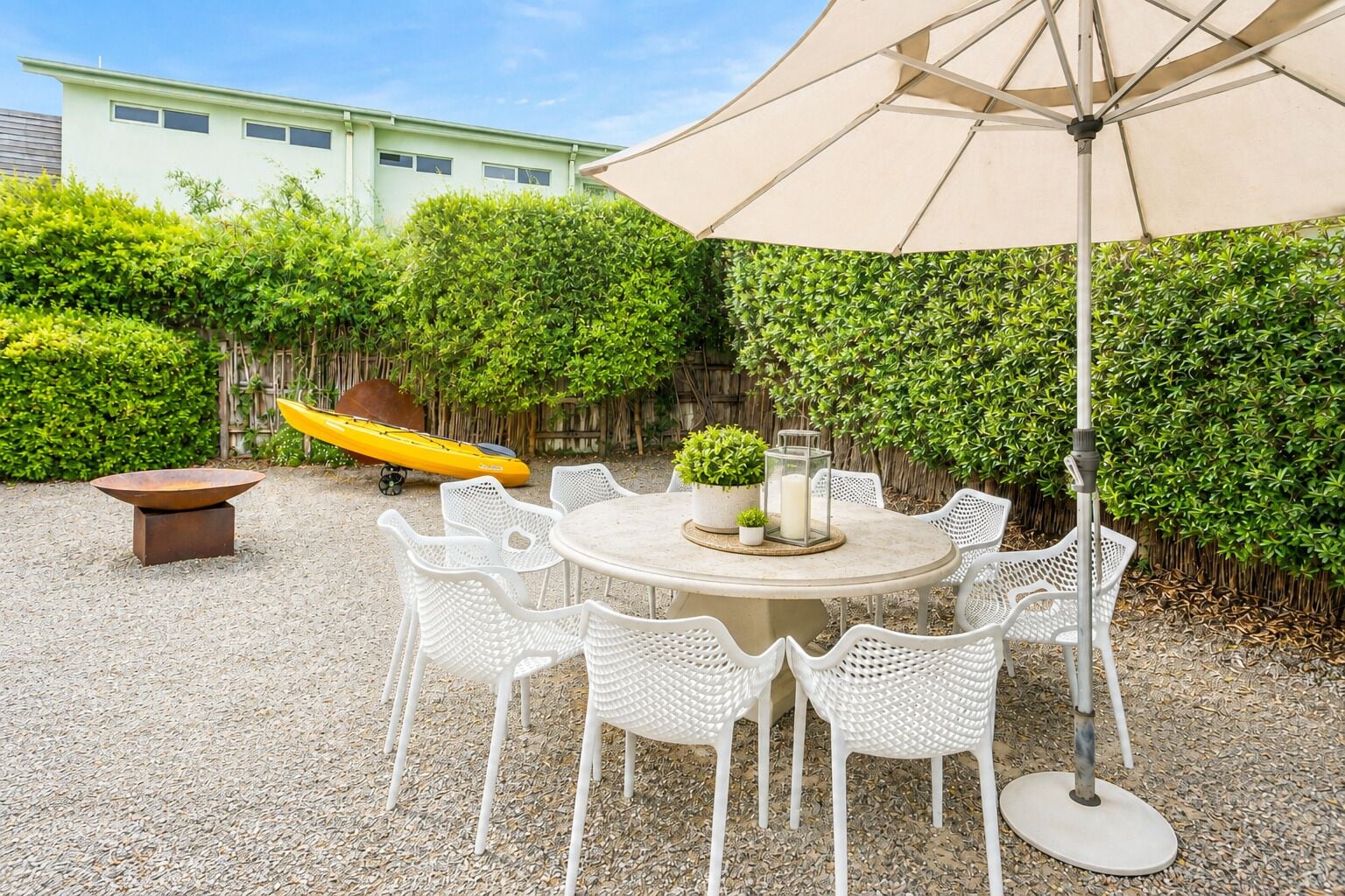 A round outdoor dining table with white chairs, a yellow kayak, and a large umbrella in a garden area.
